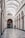 An elegant interior of an architectural hallway featuring a high arched ceiling with intricate designs. The walls are adorned with decorative molding and large arched windows. A polished patterned floor leads to a smaller archway entrance, with two people, one facing away and the other wearing a green coat, heading towards a shop named 'Librairie'. Chandeliers hang from the ceiling, adding a classic touch to the ambiance.