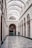 An elegant interior of an architectural hallway featuring a high arched ceiling with intricate designs. The walls are adorned with decorative molding and large arched windows. A polished patterned floor leads to a smaller archway entrance, with two people, one facing away and the other wearing a green coat, heading towards a shop named 'Librairie'. Chandeliers hang from the ceiling, adding a classic touch to the ambiance.