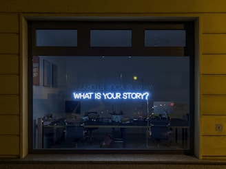 window with neon sign that says What is your story?