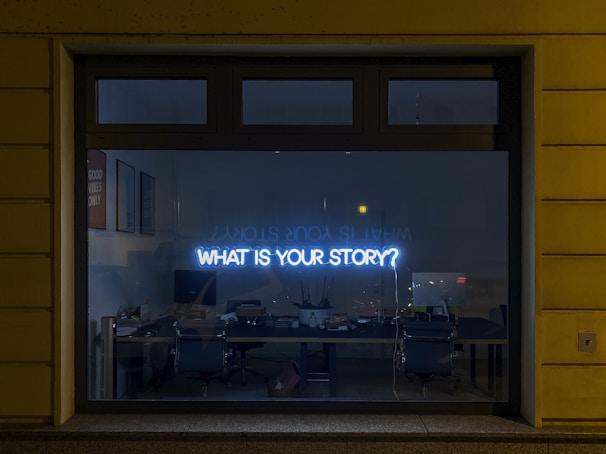 A large window reveals the interior of an office space where a neon sign with the words 'WHAT IS YOUR STORY?' is prominently displayed. The dimly lit room features several office chairs, a long desk cluttered with various items, computers, and a plant. Posters on the wall include a visible one stating 'GOOD VIBES ONLY,' contributing to the decor.