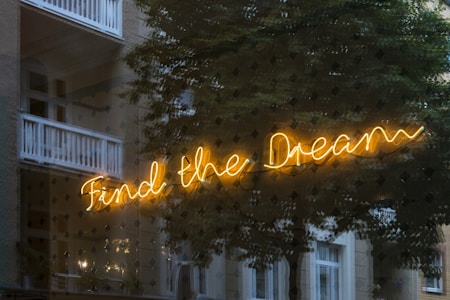 A neon sign with the phrase 'Find the Dream' is displayed against a background of a building exterior with trees partially reflected in the glass. The sign is bright yellow, contrasting with the darker, muted tones of the building and trees.