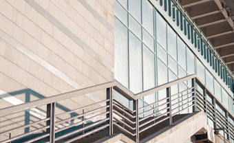 A modern architectural structure featuring a staircase with metal railings that ascends alongside a wall with horizontal lines. The wall is adjacent to a section of large reflective glass windows.