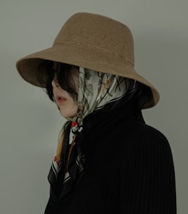 A person is wearing a brown wide-brimmed hat and a patterned scarf covering their head. Their hair is mostly hidden under the scarf, and they are dressed in a dark ribbed turtleneck sweater. The background is a muted green color, giving a vintage or artistic vibe.