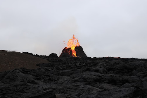 A volcanic eruption with bright orange lava spewing from its top, surrounded by dark, rugged volcanic rocks under an overcast sky.