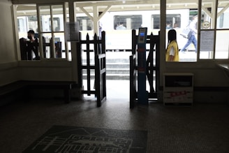 A dimly lit interior of a train station with a ticket gate prominently displayed. Outside, a train is visible, and a few people, some wearing masks, are seen on the platform. The scene is framed by windows and wooden structures, with some signs and notices visible.