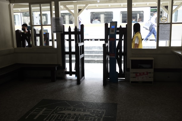 A dimly lit interior of a train station with a ticket gate prominently displayed. Outside, a train is visible, and a few people, some wearing masks, are seen on the platform. The scene is framed by windows and wooden structures, with some signs and notices visible.