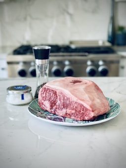 A large cut of raw marbled beef is placed on a decorative plate with a floral design, situated on a marble countertop. In the background, a pepper grinder and a small jar of salt are visible, with a modern stovetop providing a blurred backdrop.