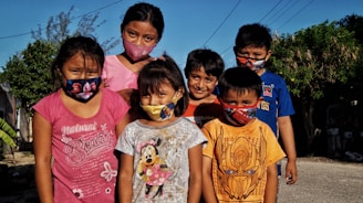 A group of six children wearing colorful masks stand outside on a sunny day, surrounded by greenery and residential buildings. They are dressed in casual clothing with various prints, and the background includes clear blue skies and power lines.