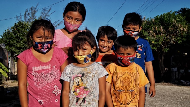 A group of six children wearing colorful masks stand outside on a sunny day, surrounded by greenery and residential buildings. They are dressed in casual clothing with various prints, and the background includes clear blue skies and power lines.