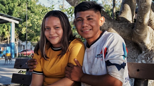 A young boy and girl are sitting closely on a bench under a tree, in a bright and sunny outdoor setting. The girl is wearing a yellow shirt while the boy is in a white shirt with colorful designs. They are smiling warmly, suggesting a joyful or affectionate moment.
