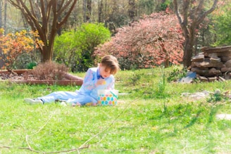 A young child is sitting on grass in a garden, focused on an Easter egg. The garden is lush with green grass and various blooming flowers and plants, including a tree with red and orange blossoms. There are stone slabs stacked to the side.