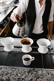 A person is pouring hot water from a stainless steel kettle into a coffee filter. The setup includes three white ceramic pour-over coffee drippers, with one being actively used to brew coffee, which is dripping into a cup below. The individual is wearing a white shirt and a black vest, suggesting a professional coffee-making setting, likely in a cafe or a coffee shop.