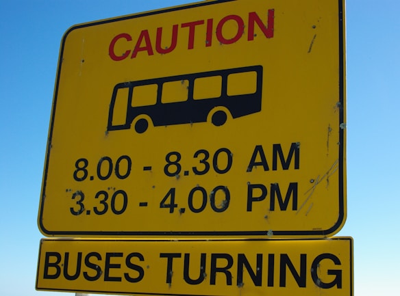 A yellow caution sign with a black silhouette of a bus indicates specific times: 8.00 - 8.30 AM and 3.30 - 4.00 PM. Below, another sign in yellow states 'BUSES TURNING'. The signs are against a clear blue sky background.