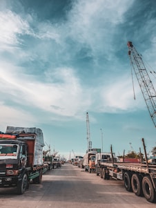 A bustling port scene featuring numerous trucks lined up along a concrete road. Cranes tower above, indicating active shipping and cargo operations. The sky is partly cloudy, casting soft shadows on the ground.