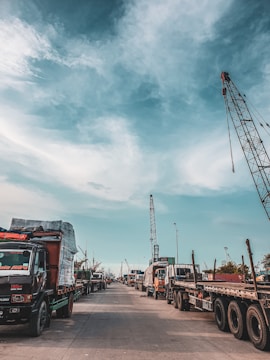 A bustling port scene featuring numerous trucks lined up along a concrete road. Cranes tower above, indicating active shipping and cargo operations. The sky is partly cloudy, casting soft shadows on the ground.