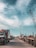A bustling port scene featuring numerous trucks lined up along a concrete road. Cranes tower above, indicating active shipping and cargo operations. The sky is partly cloudy, casting soft shadows on the ground.