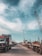 A bustling port scene featuring numerous trucks lined up along a concrete road. Cranes tower above, indicating active shipping and cargo operations. The sky is partly cloudy, casting soft shadows on the ground.