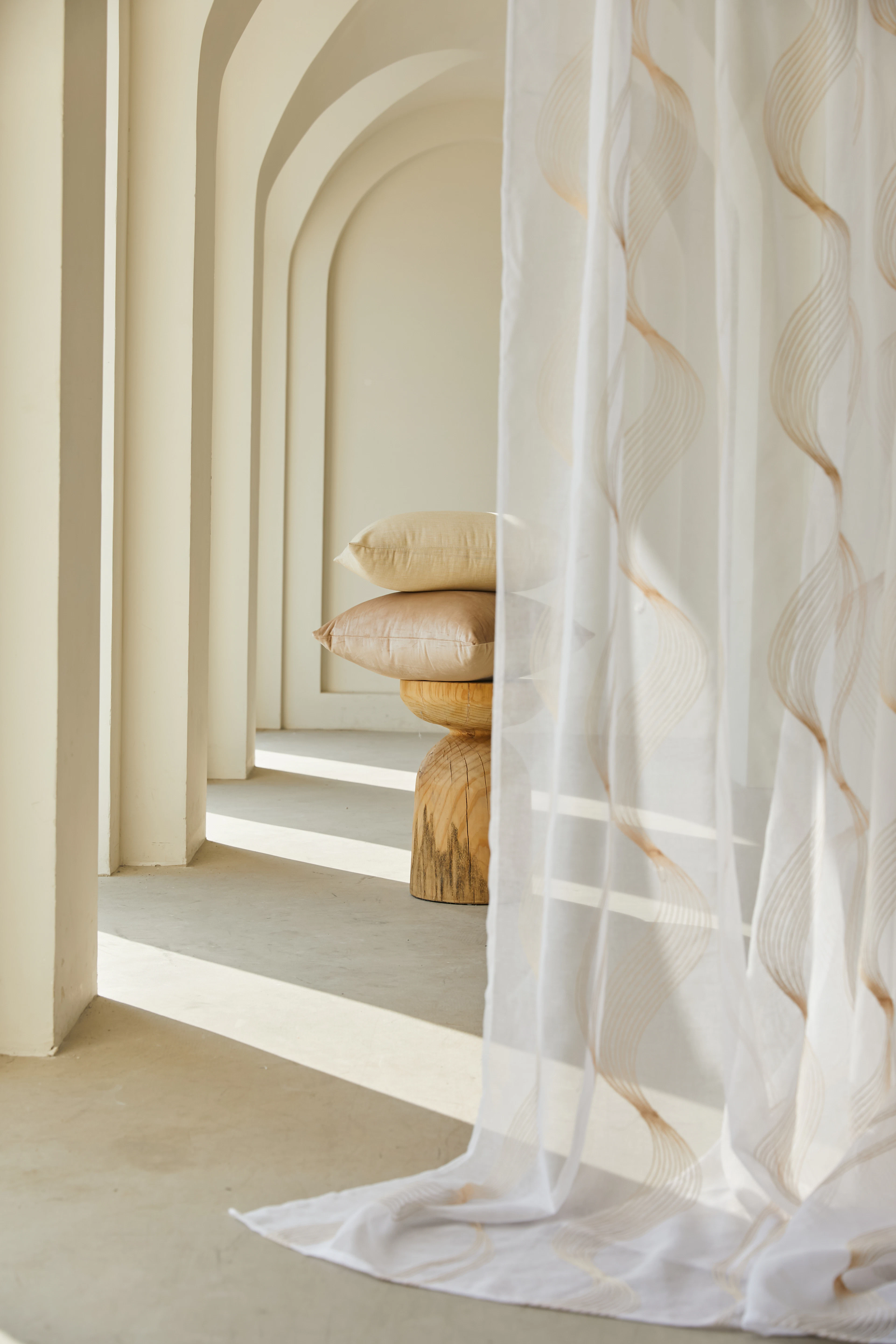 Receding plaster arches washed with daylight, a sheer linen curtain and a carved timber stool