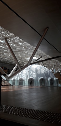 A modern architectural space featuring a large, white dome with multiple arched windows. The ceiling has an intricate lattice design, allowing natural light to filter through, creating dynamic patterns of light and shadow. Thick, metallic beams converge towards the dome, contributing to the contemporary aesthetic.