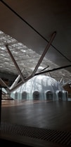 A modern architectural space featuring a large, white dome with multiple arched windows. The ceiling has an intricate lattice design, allowing natural light to filter through, creating dynamic patterns of light and shadow. Thick, metallic beams converge towards the dome, contributing to the contemporary aesthetic.