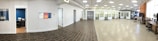 A spacious office interior with a tiled floor transitioning to a polished concrete surface. There are several closed office doors along the left side of the room, some with small windows. Office furniture is arranged around the room, including desks and chairs. A small office room is visible through an open door, equipped with a desk and chair. The ceiling features a grid of overhead lights, providing a well-lit atmosphere.