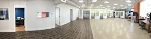 A spacious office interior with a tiled floor transitioning to a polished concrete surface. There are several closed office doors along the left side of the room, some with small windows. Office furniture is arranged around the room, including desks and chairs. A small office room is visible through an open door, equipped with a desk and chair. The ceiling features a grid of overhead lights, providing a well-lit atmosphere.
