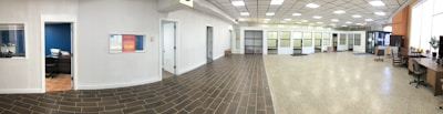 A spacious office interior with a tiled floor transitioning to a polished concrete surface. There are several closed office doors along the left side of the room, some with small windows. Office furniture is arranged around the room, including desks and chairs. A small office room is visible through an open door, equipped with a desk and chair. The ceiling features a grid of overhead lights, providing a well-lit atmosphere.