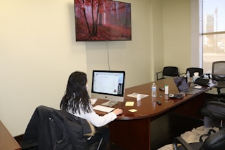 A person with long hair is seated at a large wooden desk, working on a desktop computer. There is a water bottle, papers, sticky notes, and electronics such as a laptop and a remote control on the table. The room is a conference space with several office chairs. A large screen TV on the wall displays an image of a forest with red foliage.