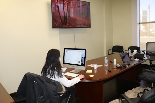 A person with long hair is seated at a large wooden desk, working on a desktop computer. There is a water bottle, papers, sticky notes, and electronics such as a laptop and a remote control on the table. The room is a conference space with several office chairs. A large screen TV on the wall displays an image of a forest with red foliage.