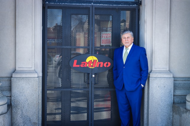 A man in a blue suit stands in front of a metal gate with the word 'Latino' on a sign. The gate is flanked by stone columns. The man has grey hair and wears a light green tie, exuding a formal appearance.
