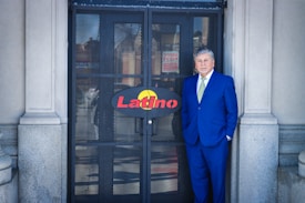A man in a blue suit stands in front of a metal gate with the word 'Latino' on a sign. The gate is flanked by stone columns. The man has grey hair and wears a light green tie, exuding a formal appearance.