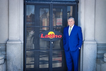 A man in a blue suit stands in front of a metal gate with the word 'Latino' on a sign. The gate is flanked by stone columns. The man has grey hair and wears a light green tie, exuding a formal appearance.