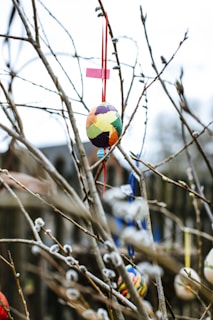Colorful Easter eggs hang from the branches of a budding tree, decorated with ribbons. Some branches have small, soft buds, suggesting the early stages of spring.