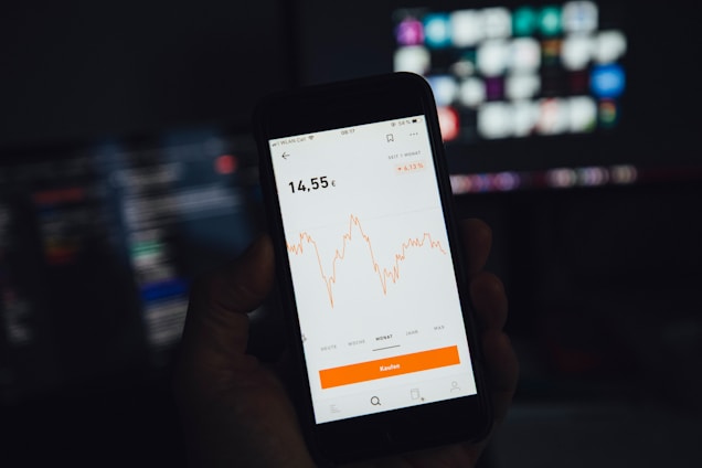 A hand is holding a smartphone displaying a financial chart with fluctuations in orange lines. The background features blurred computer screens with a dark theme.