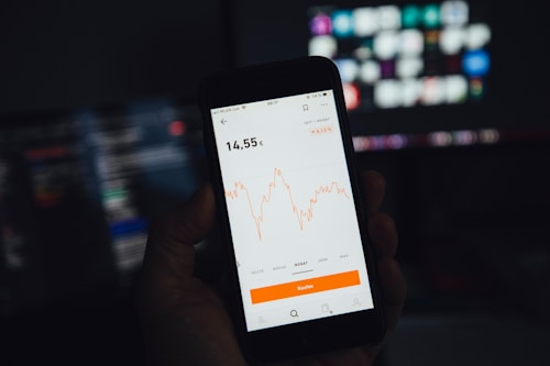 A hand is holding a smartphone displaying a financial chart with fluctuations in orange lines. The background features blurred computer screens with a dark theme.