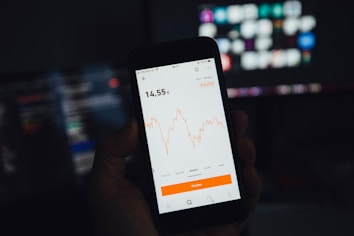 A hand is holding a smartphone displaying a financial chart with fluctuations in orange lines. The background features blurred computer screens with a dark theme.