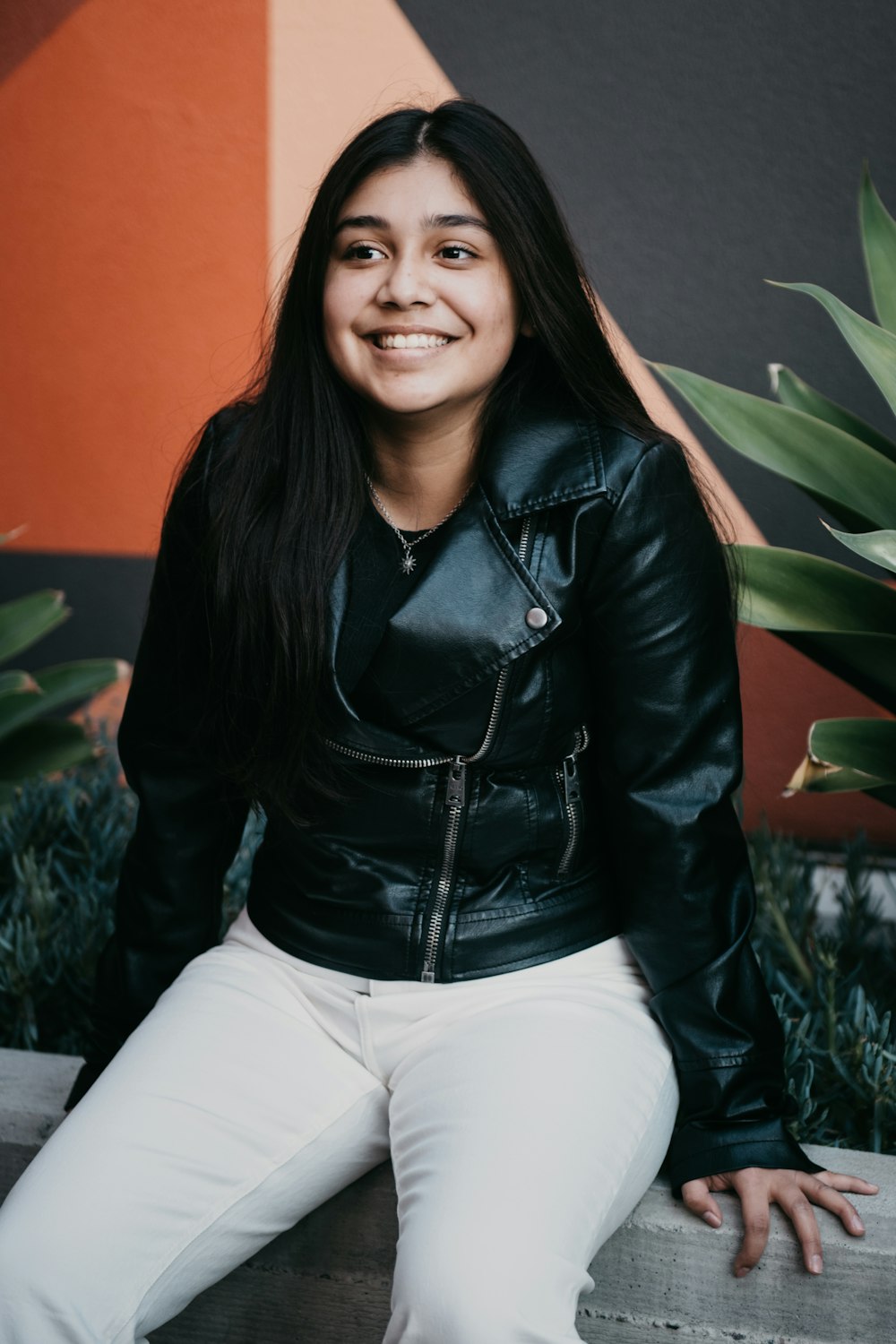 woman in black leather jacket and white pants sitting on white textile