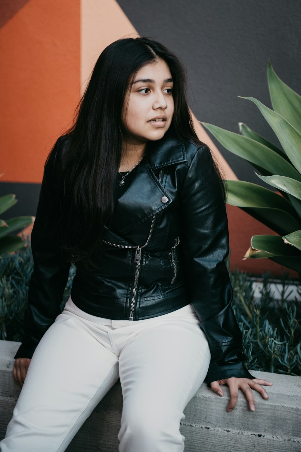 woman in black leather jacket and white pants sitting on white bed
