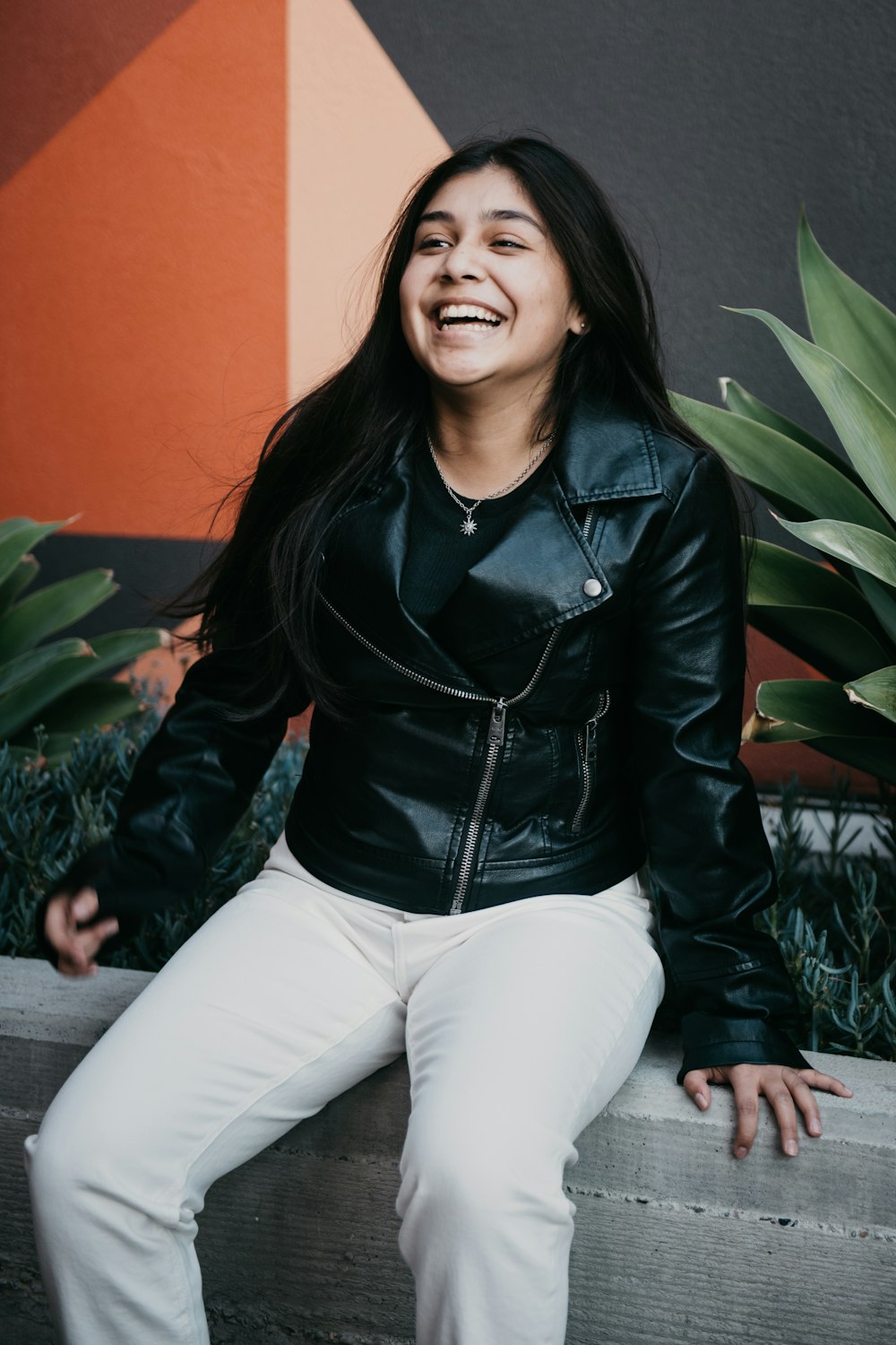 woman in black leather jacket and white pants sitting on green plant