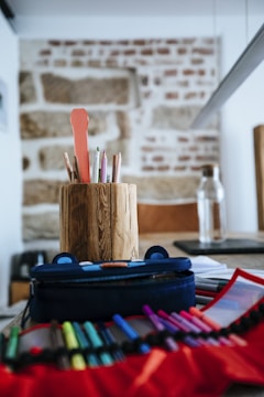 A collection of colored pens and pencils organized in a rustic, cylindrical wooden holder. The scene takes place on a table with additional stationery items including a pencil case and various writing instruments spread out in the foreground. In the background, there is a stone brick wall and a transparent bottle.
