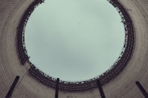 A view from the inside of a large industrial cooling tower, looking up towards the sky. The structure is cylindrical with a wide opening at the top, displaying a grid-like pattern of metal beams around the rim. The interior walls are made of concrete with a slightly weathered appearance.