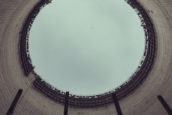A view from the inside of a large industrial cooling tower, looking up towards the sky. The structure is cylindrical with a wide opening at the top, displaying a grid-like pattern of metal beams around the rim. The interior walls are made of concrete with a slightly weathered appearance.