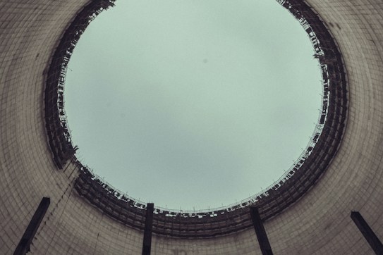 A view from the inside of a large industrial cooling tower, looking up towards the sky. The structure is cylindrical with a wide opening at the top, displaying a grid-like pattern of metal beams around the rim. The interior walls are made of concrete with a slightly weathered appearance.
