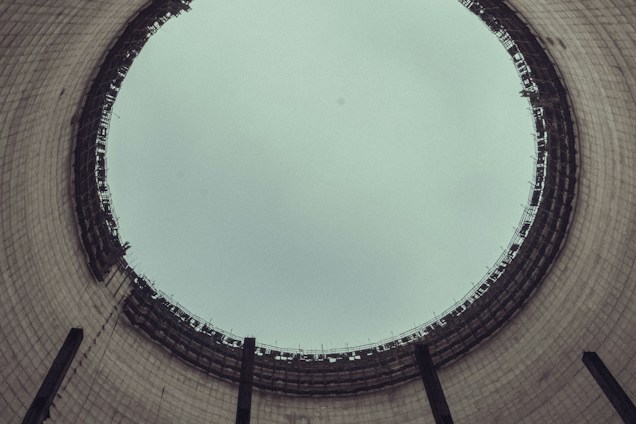 A view from the inside of a large industrial cooling tower, looking up towards the sky. The structure is cylindrical with a wide opening at the top, displaying a grid-like pattern of metal beams around the rim. The interior walls are made of concrete with a slightly weathered appearance.