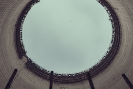 A view from the inside of a large industrial cooling tower, looking up towards the sky. The structure is cylindrical with a wide opening at the top, displaying a grid-like pattern of metal beams around the rim. The interior walls are made of concrete with a slightly weathered appearance.