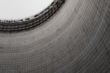 A large, curved concrete structure with a grid-like pattern and an unfinished top with scaffolding. The texture of the concrete and the expansive form create an industrial look.