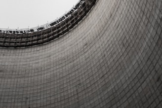 A large, curved concrete structure with a grid-like pattern and an unfinished top with scaffolding. The texture of the concrete and the expansive form create an industrial look.