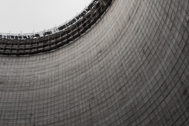 A large, curved concrete structure with a grid-like pattern and an unfinished top with scaffolding. The texture of the concrete and the expansive form create an industrial look.