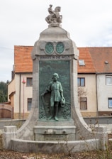A monument with a detailed bronze relief depicting a young figure holding a staff or walking stick. Above the relief is a sculpted figure, possibly a child riding a creature. The monument features inscriptions and is situated in front of a building with a tiled roof.