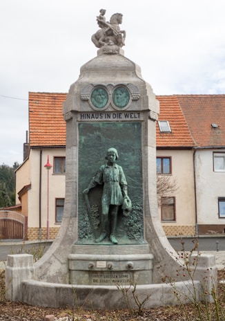 A monument with a detailed bronze relief depicting a young figure holding a staff or walking stick. Above the relief is a sculpted figure, possibly a child riding a creature. The monument features inscriptions and is situated in front of a building with a tiled roof.